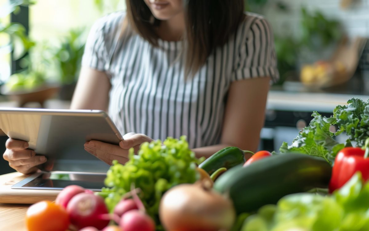 Woman using a tablet in a kitchen filled with fresh vegetables. Concept of meal planning and healthy eating.